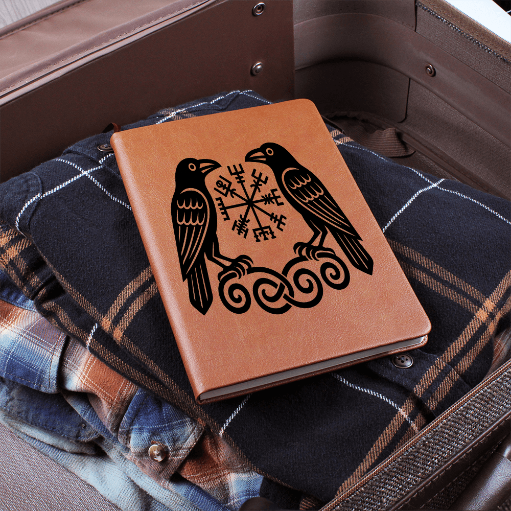 Tan leather journal featuring two Norse ravens and a Vegvisir rune compass over knotwork, resting on folded plaid clothing inside an open suitcase for travel.