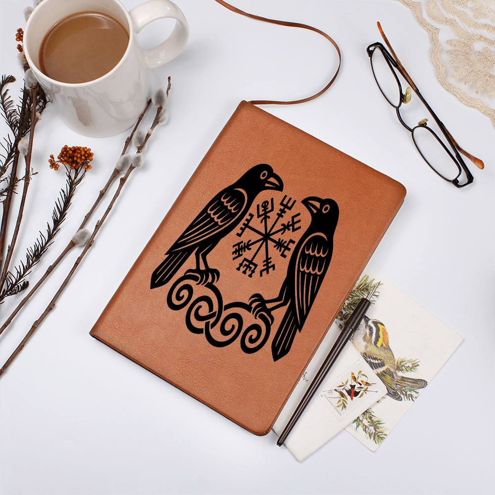 Tan leather journal with black Norse raven design flanking a Vegvisir rune compass on knotwork, shown on a white desk beside coffee mug, eyeglasses, twigs, pen, and bird-illustration postcards.