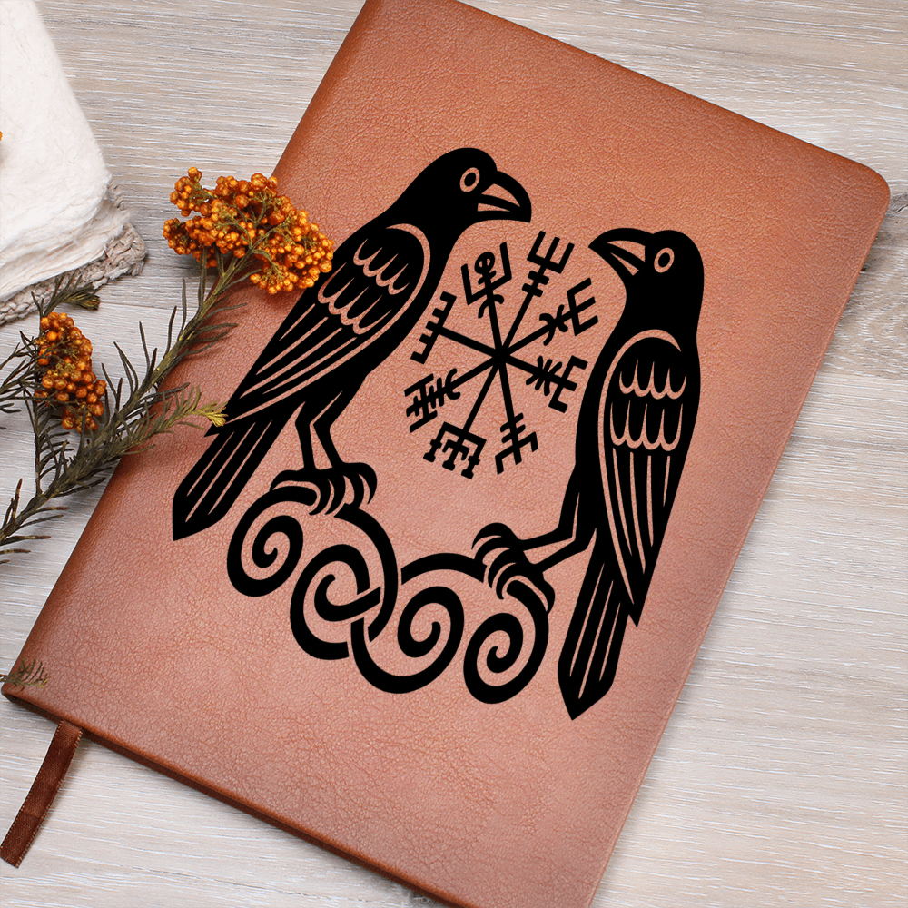 Brown leather journal featuring black Viking ravens (Huginn and Muninn) flanking a Vegvisir rune symbol on a wood desk, ribbon bookmark, orange berry sprigs.