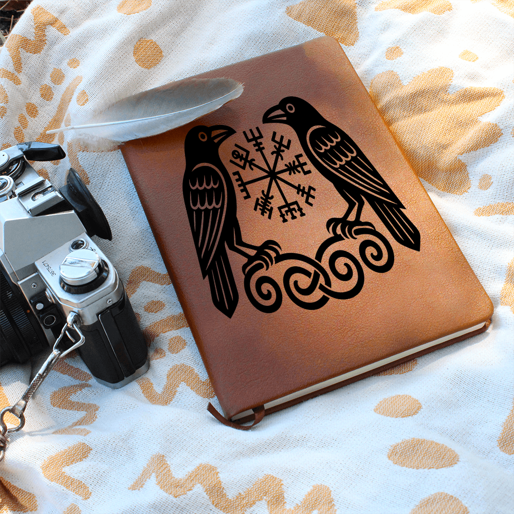 Brown leather journal featuring two Viking ravens flanking a Vegvisir rune symbol on knotwork, resting on patterned cloth beside a white feather and a vintage film camera.