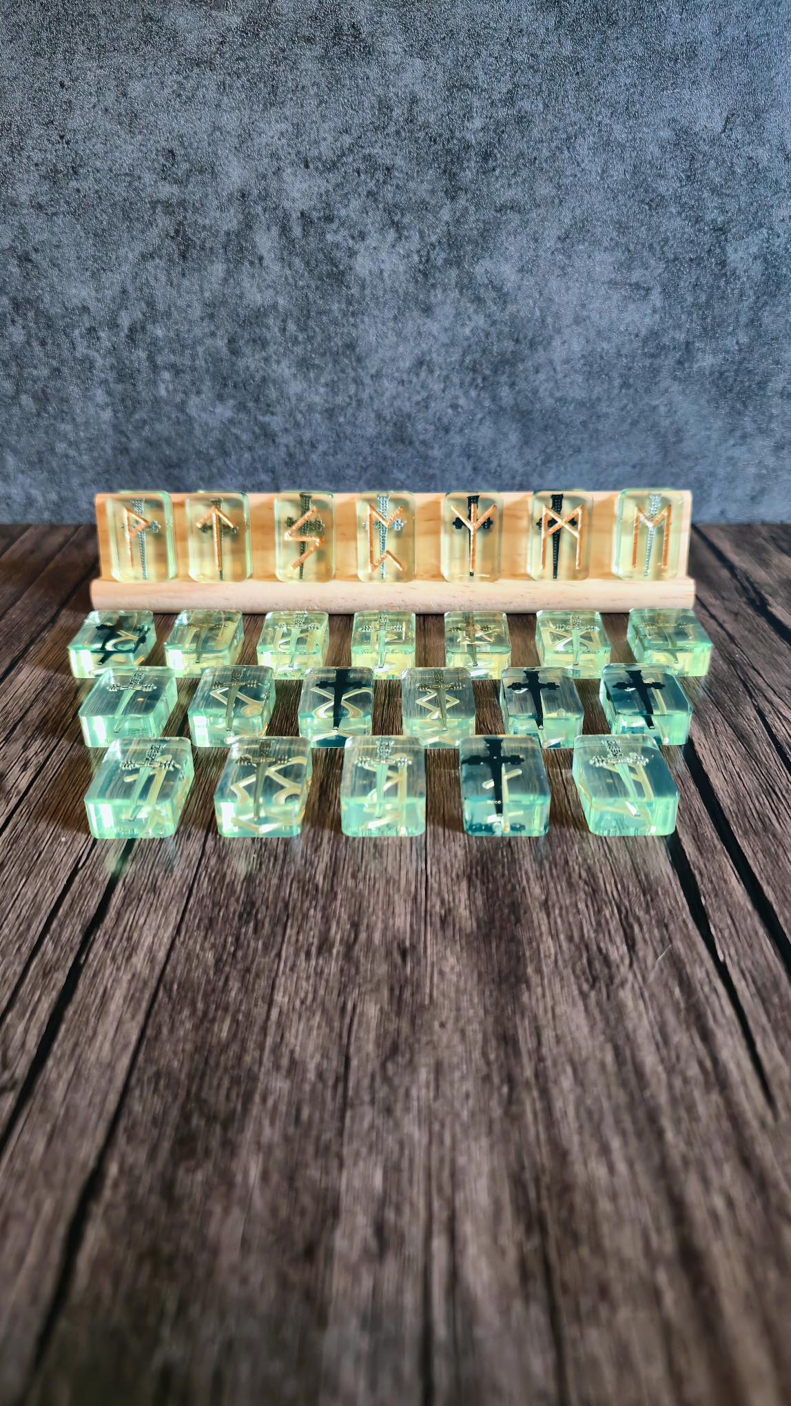 Greenish clear runestones with swords embedded with gold runic symbols on birchwood stand with grey background on wooden table.