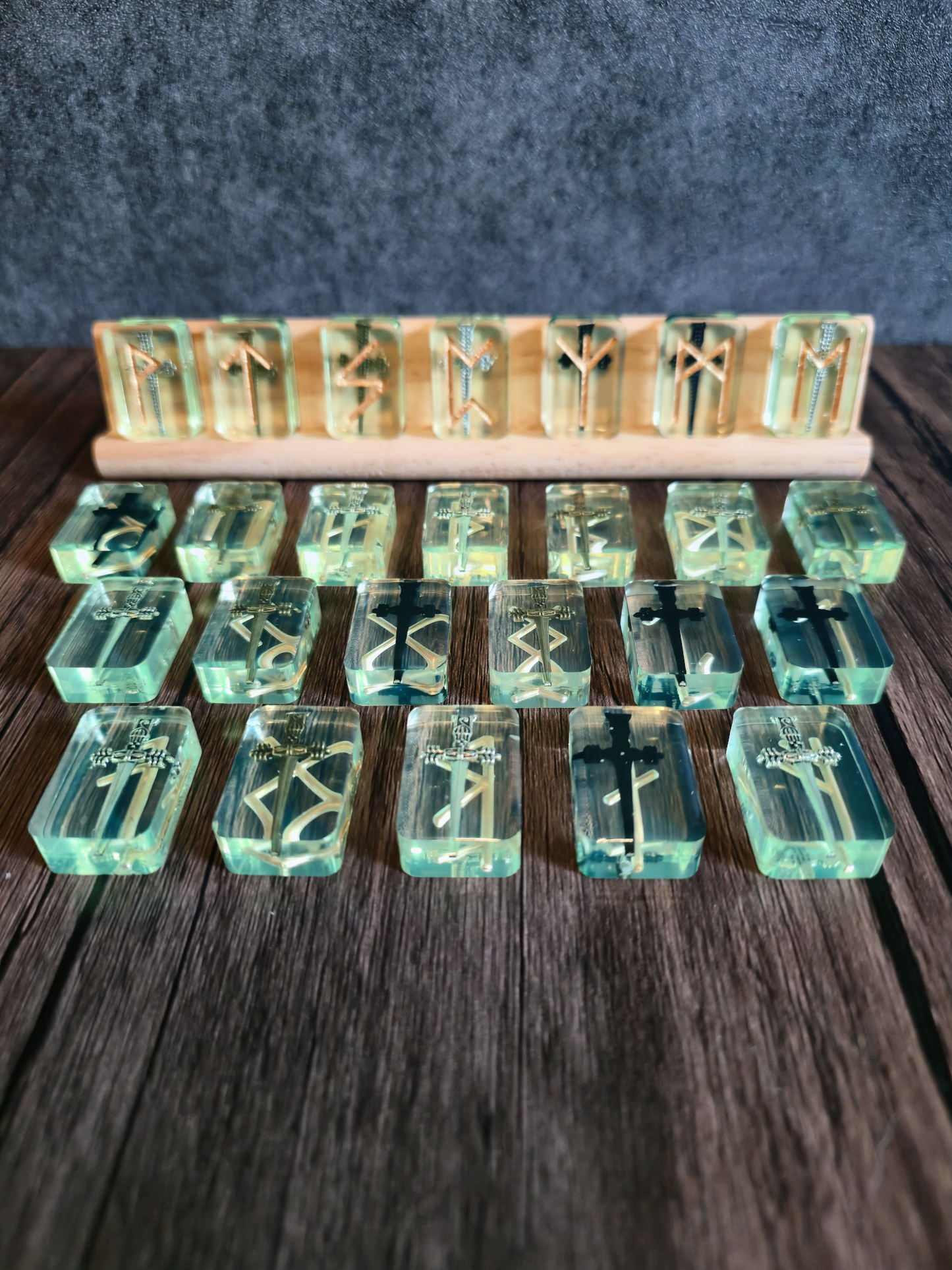 Greenish clear runestones with swords embedded with gold runic symbols on birchwood stand with grey background on wooden table.