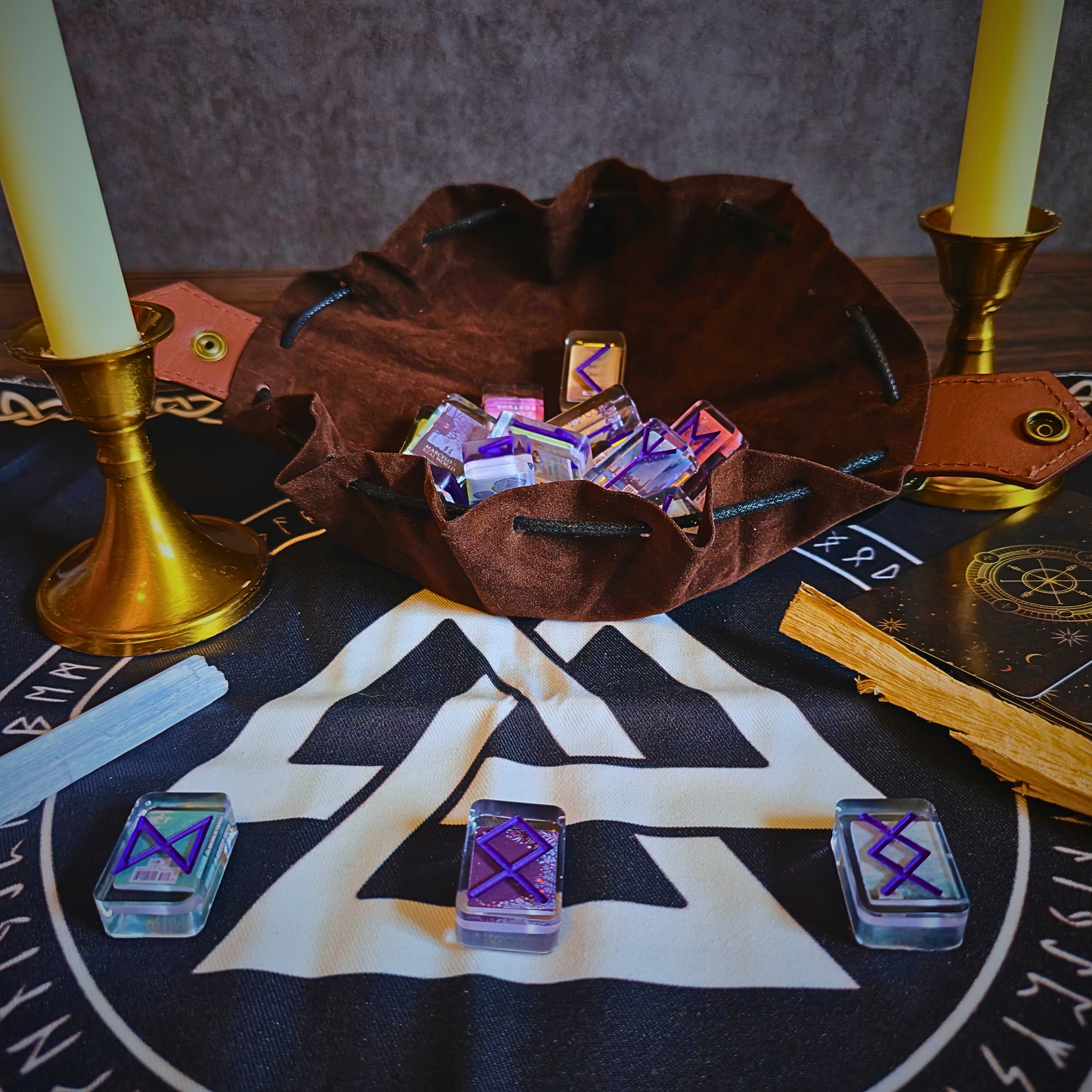 A rustic brown suede drawstring pouch filled with translucent glass runestones engraved in purple Norse runes, arranged atop a black-and-white Viking altar cloth, with brass candle holders, a selenite wand on the left, and a palo santo stick on the right.