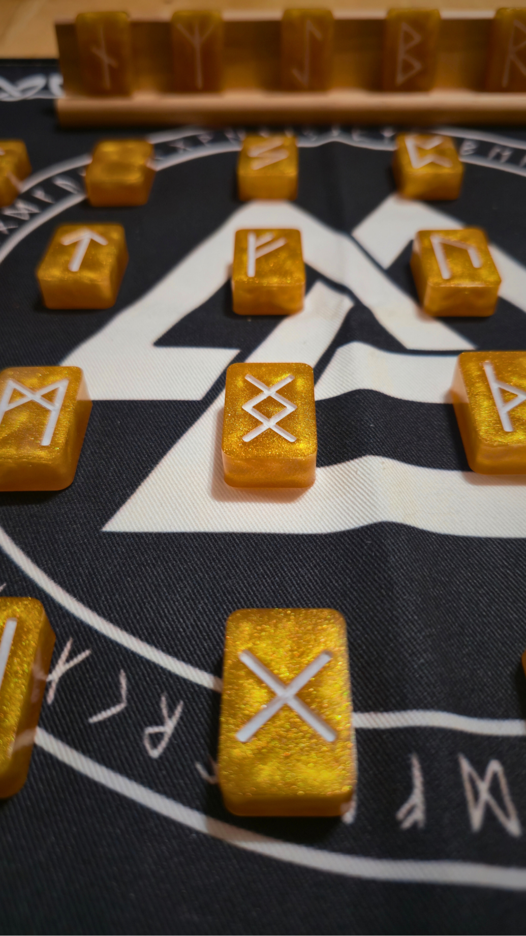 Asgardian Gold Ingot Runestone Set: close-up of shimmering gold resin rune ingots engraved with white Elder Futhark symbols, arranged on a black rune cloth printed with a white Valknut and circular runic border, with a birch wood display stand in the background.