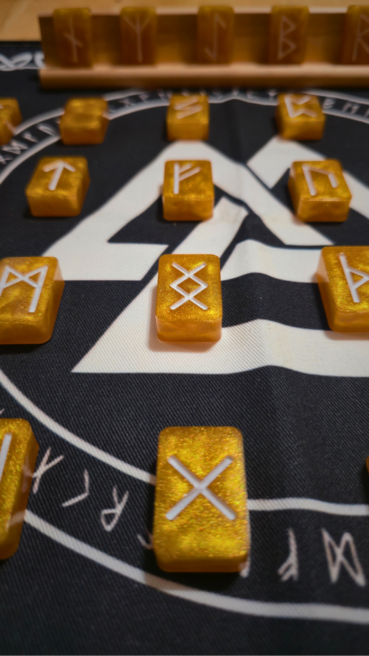 Asgardian Gold Ingot Runestone Set: close-up of shimmering gold resin rune ingots engraved with white Elder Futhark symbols, arranged on a black rune cloth printed with a white Valknut and circular runic border, with a birch wood display stand in the background.