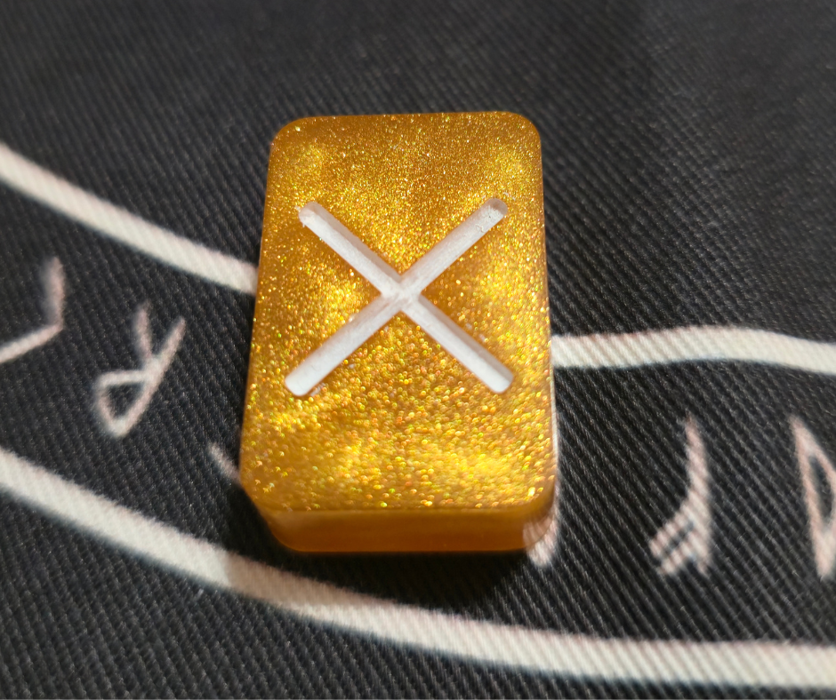 Close-up of a glittering gold resin runestone ingot from the Asgardian Gold Ingot Runestone Set, etched with a white Gebo (X) Elder Futhark rune, resting on a black cloth printed with runic script.