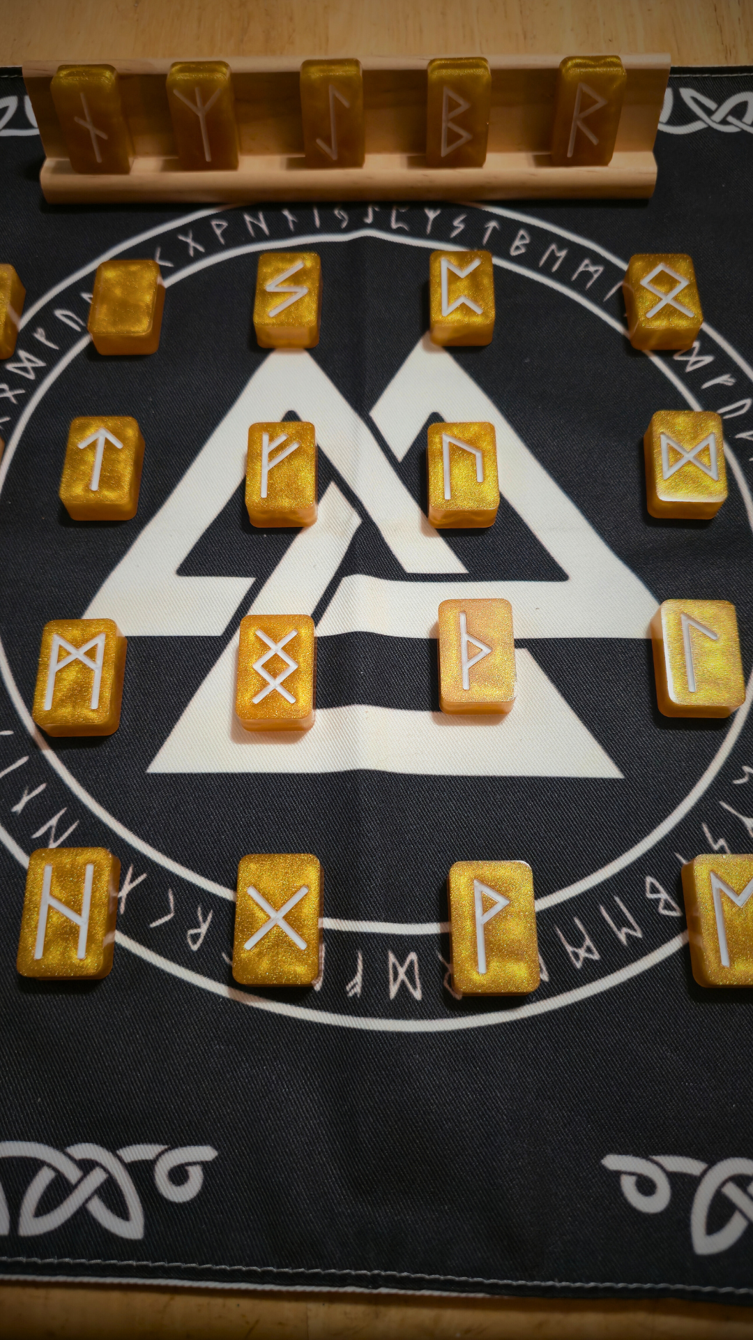 Asgardian Gold Ingot Runestone Set: 24 shimmering gold resin Elder Futhark runes with crisp white symbols arranged in a circle on a black rune cloth featuring a white Valknut design, with five rune ingots displayed on a birch wood stand above.