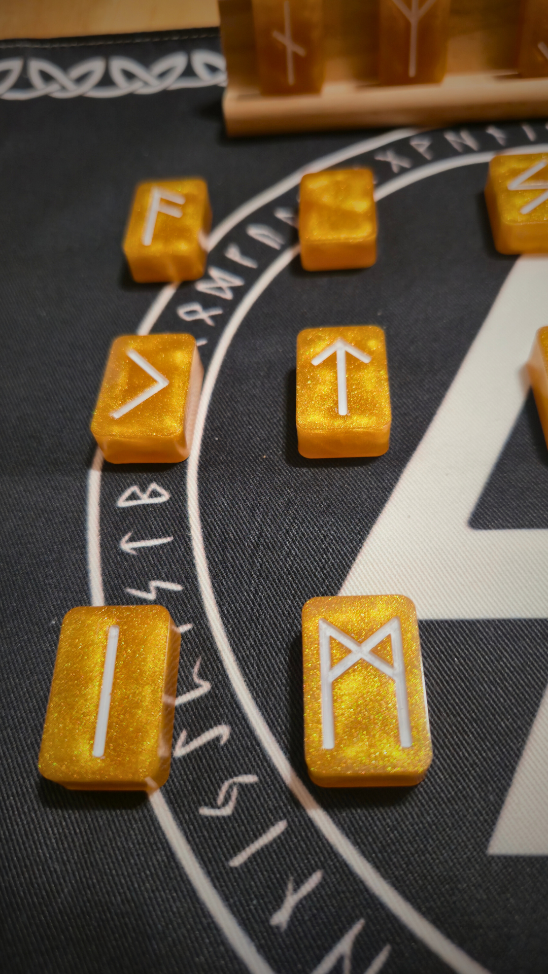 Shimmering gold resin runestones etched with white Elder Futhark runes, arranged on a black cloth printed with a white Valknut symbol and circular runic border, with a wooden display stand of additional ingots behind them.