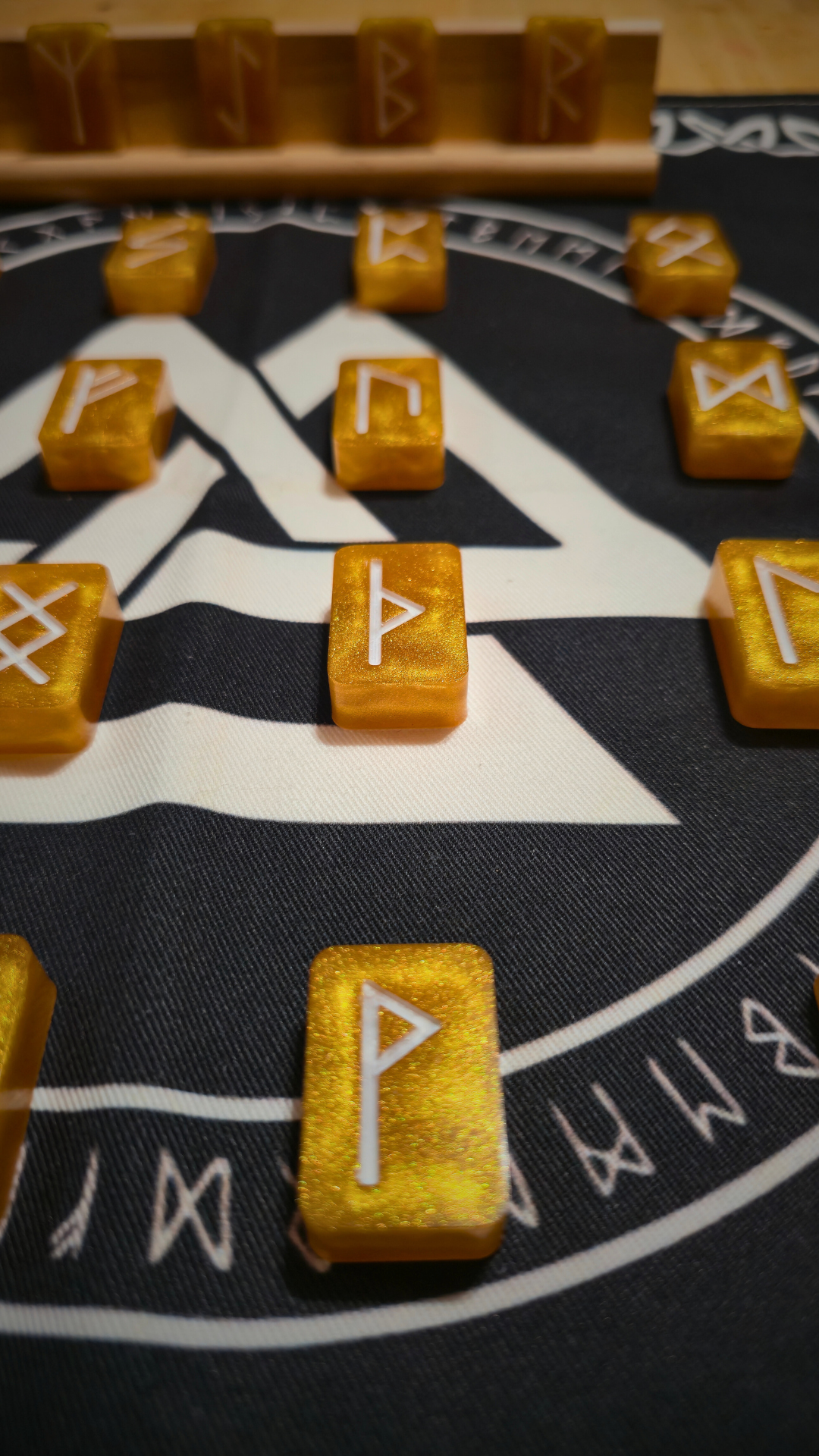 Close-up of a shimmering gold resin runestone ingot from the Asgardian Gold Ingot Runestone Set, etched with a white Elder Futhark Perthro (ᛈ) rune, resting on a black cloth printed with a white Valknut symbol and circular runic border.