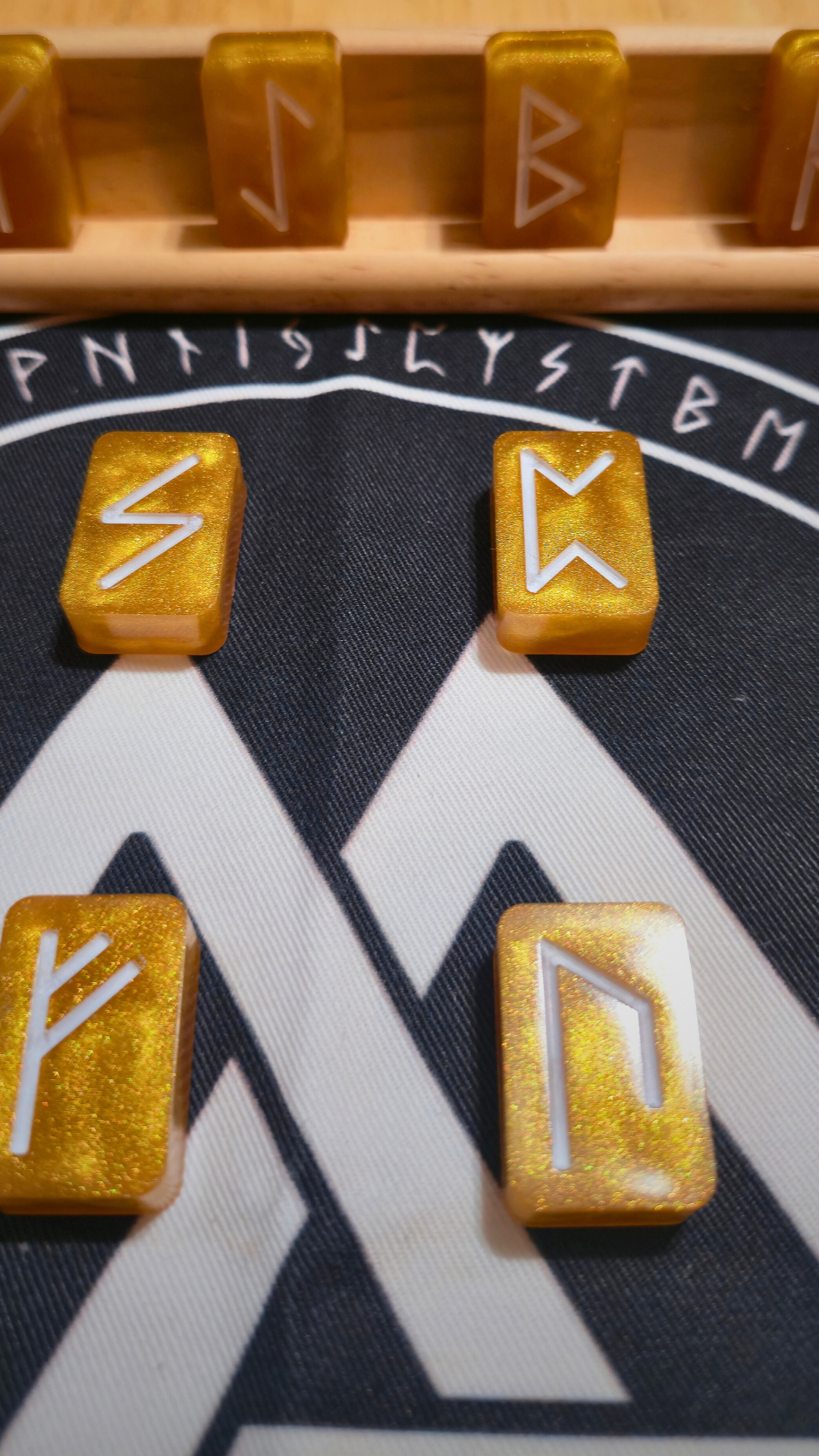 Close-up of shimmering gold resin runestone ingots from the Asgardian Gold Ingot Runestone Set, each etched with a white Elder Futhark rune, resting on a black rune cloth printed with a white Valknut and circular runic border, with a wooden display rack of additional ingots in the background.