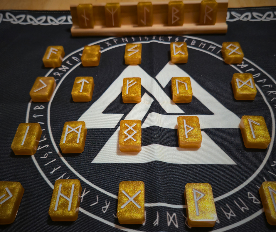 Asgardian Gold Ingot Runestone Set: 24 shimmering gold resin runes with crisp white Elder Futhark symbols arranged in a circle on a black rune cloth with a printed Valknut design, and a wooden display stand holding five rune ingots in the background.