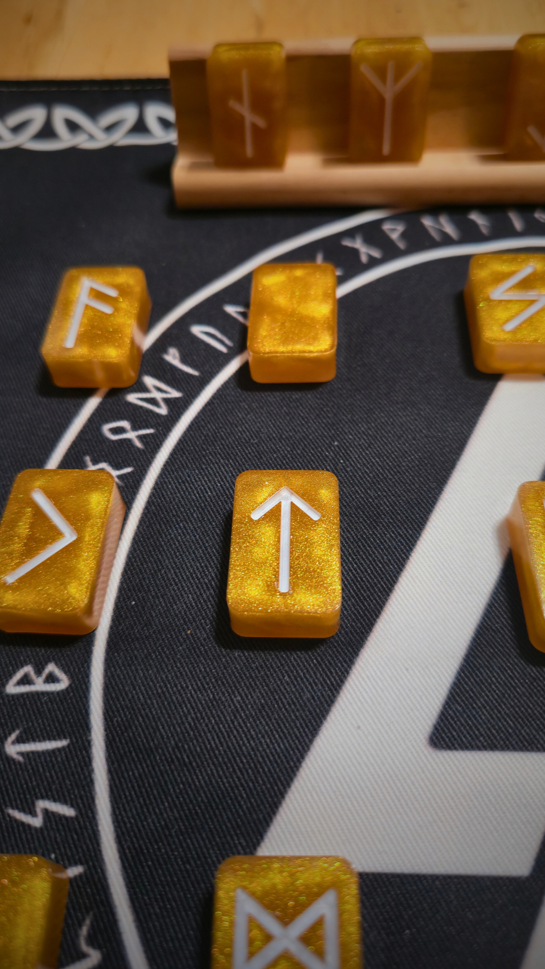 Close-up of a gold resin runestone ingot from the Asgardian Gold Ingot Runestone Set, etched with the white Tiwaz (arrow) Elder Futhark rune, resting on a black rune cloth printed with a Valknut symbol and circular runic border.