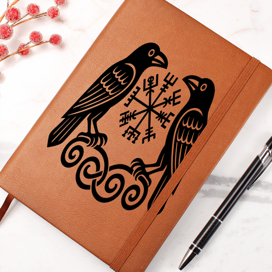 Brown leather journal featuring black Viking ravens (Huginn and Muninn) flanking a Vegvisir rune symbol, elastic strap, pen on marble, red berry sprig.
