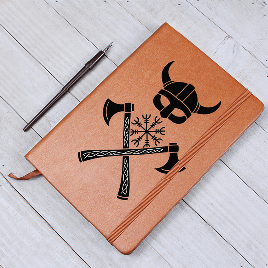 Tan vegan-leather journal on a white wood desk, featuring a black horned Viking helmet, Vegvisir rune compass, and two crossed knotwork axes; elastic closure and ribbon bookmark visible, pen nearby.
