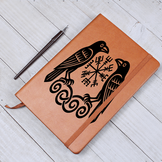 Brown leather journal featuring a black Viking design of Huginn and Muninn ravens flanking a Vegvisir rune symbol, elastic strap closed, fountain pen beside on a white wooden table.
