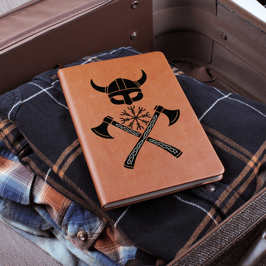 Tan vegan leather Viking journal resting on folded flannel in a suitcase, featuring a black horned helmet above a Vegvisir rune compass and crossed axes knotwork.