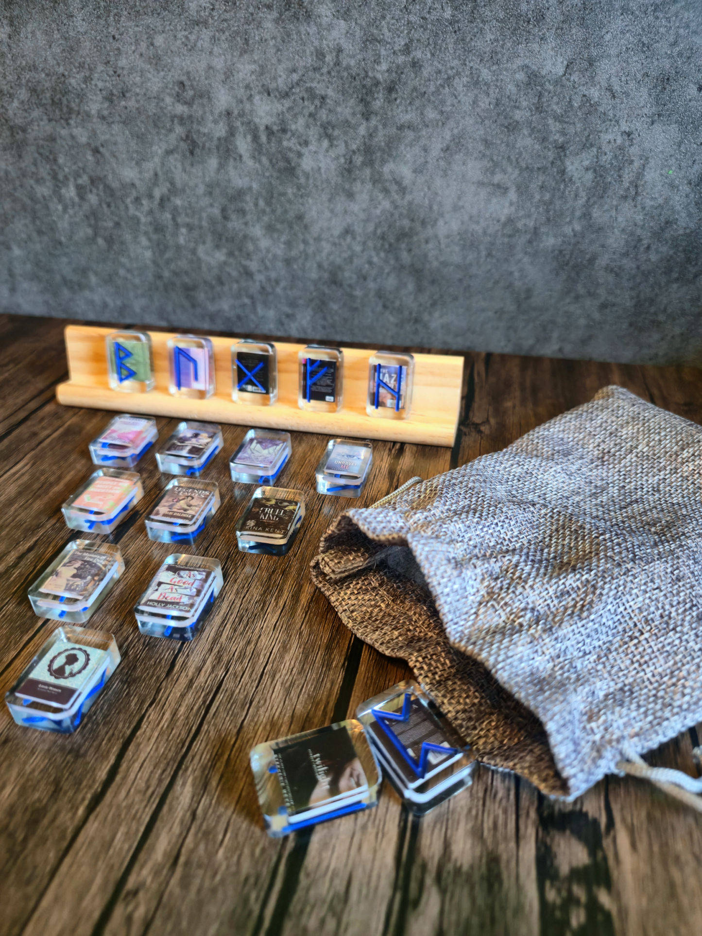 A collection of small acrylic “mini-book” rune tiles scattered on a wooden table—twelve laid out beside a woven linen drawstring pouch, and six more mounted upright on a simple wooden stand, each featuring a different cobalt-blue rune design under clear acrylic.