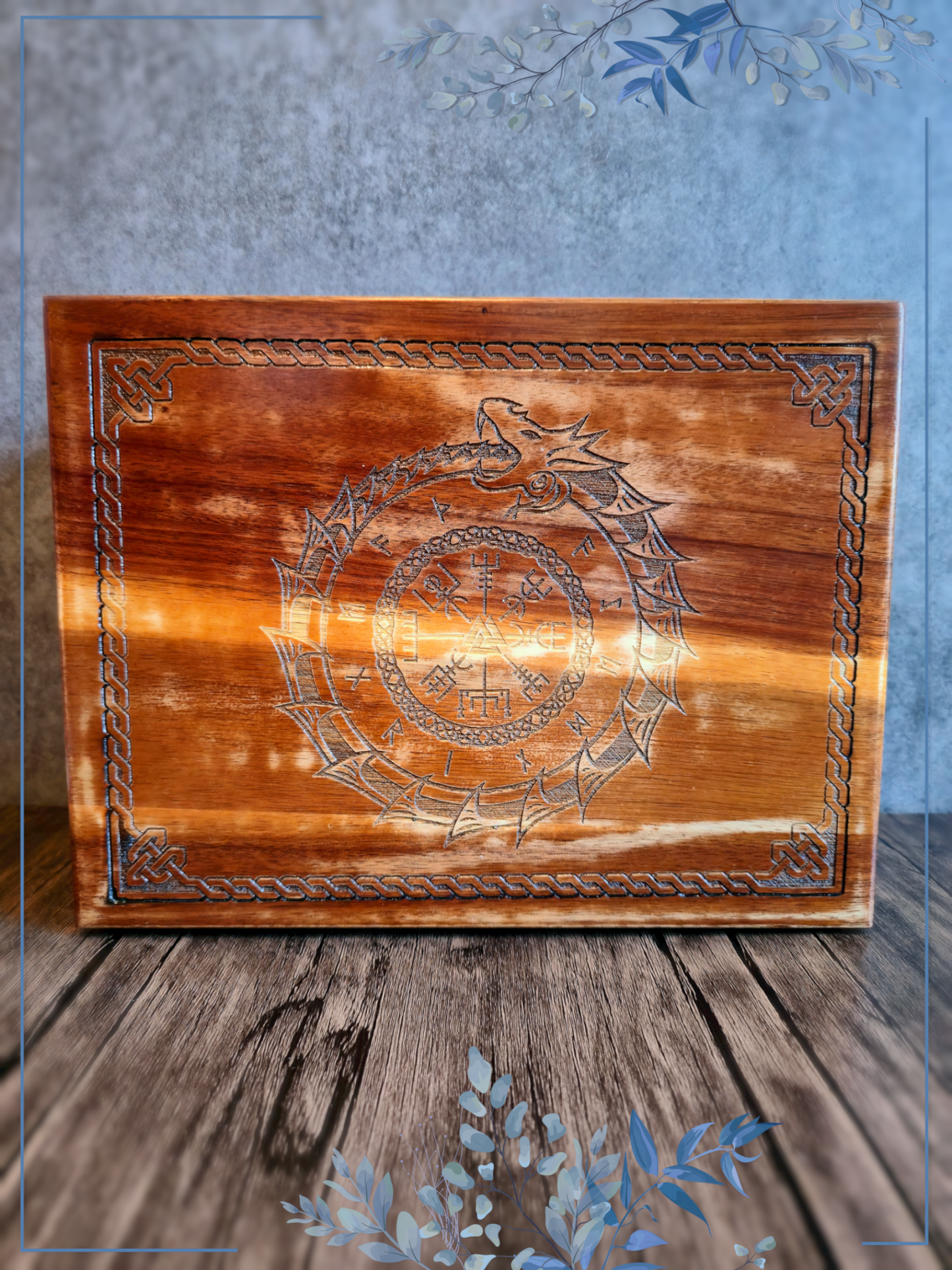 A handcrafted wooden box lid engraved with a Viking-inspired knotwork border framing a central Norse ouroboros serpent encircling a Valknut and runic symbols, set against a rustic wood grain backdrop.