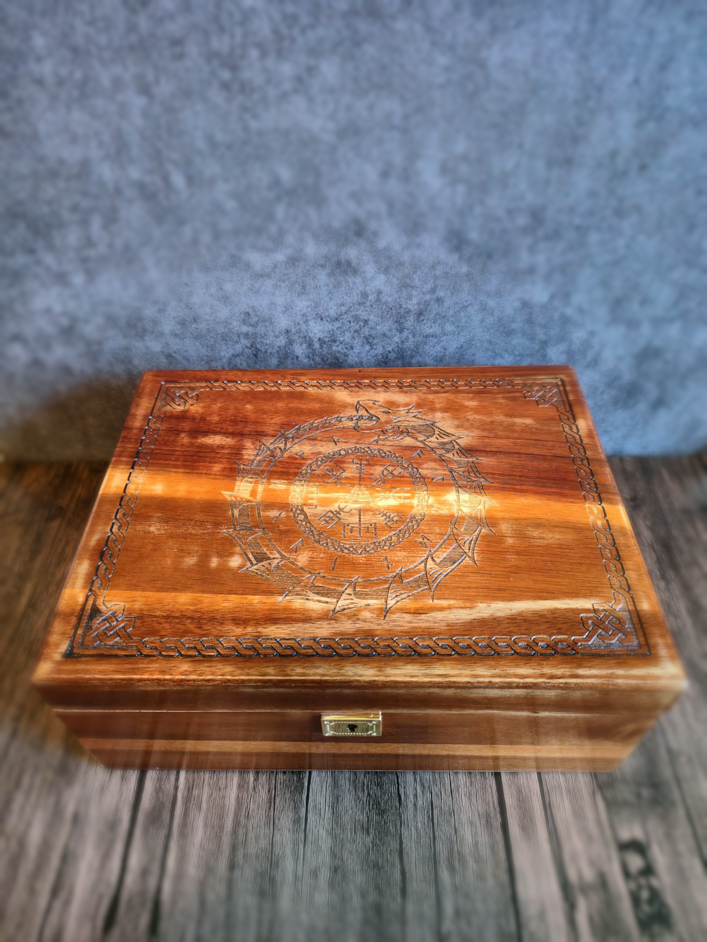 A handcrafted wooden box shown closed on a wood-grain surface, its lid engraved with an intricate Viking knotwork border and a central ouroboros serpent encircling runic symbols and a Valknut, bathed in soft, natural light.