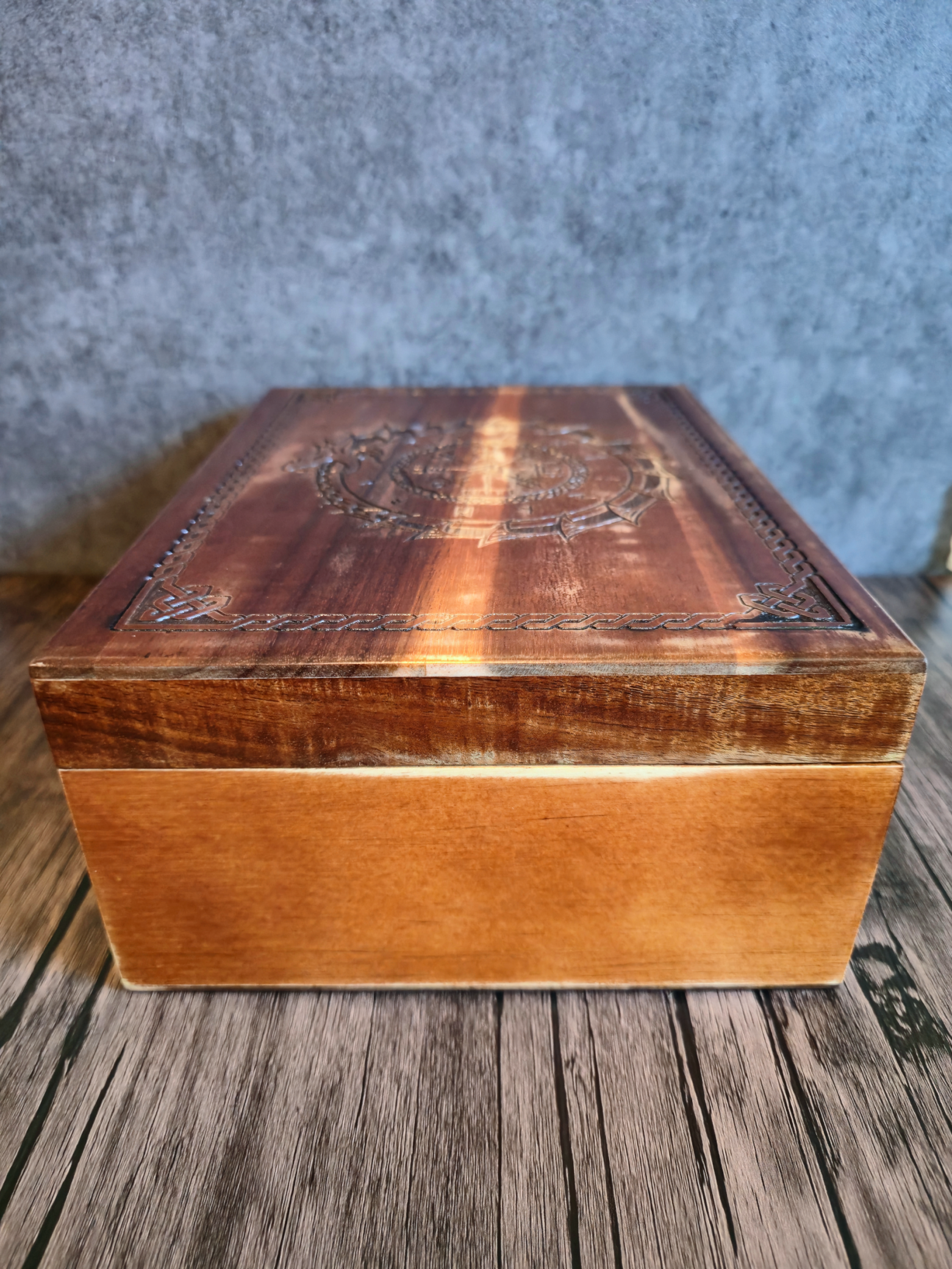 Angled side view of a closed handcrafted wooden box featuring a carved Norse dragon biting its own tail around a circle of runic symbols. The smooth stained wood grain and Celtic knotwork border are visible.