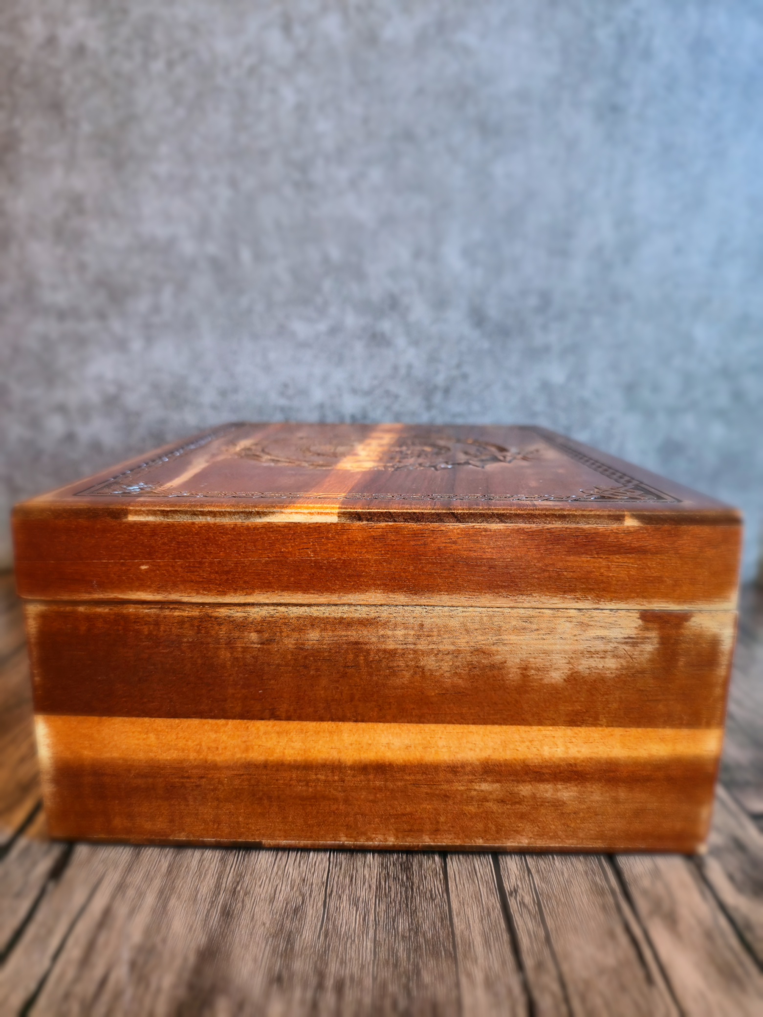 A low-angle side view of a handcrafted wooden box showing rich brown and honey-toned wood grain on its front panel, with the lid’s intricate Norse dragon-and-runic carving barely visible at the top edge against a neutral gray backdrop.