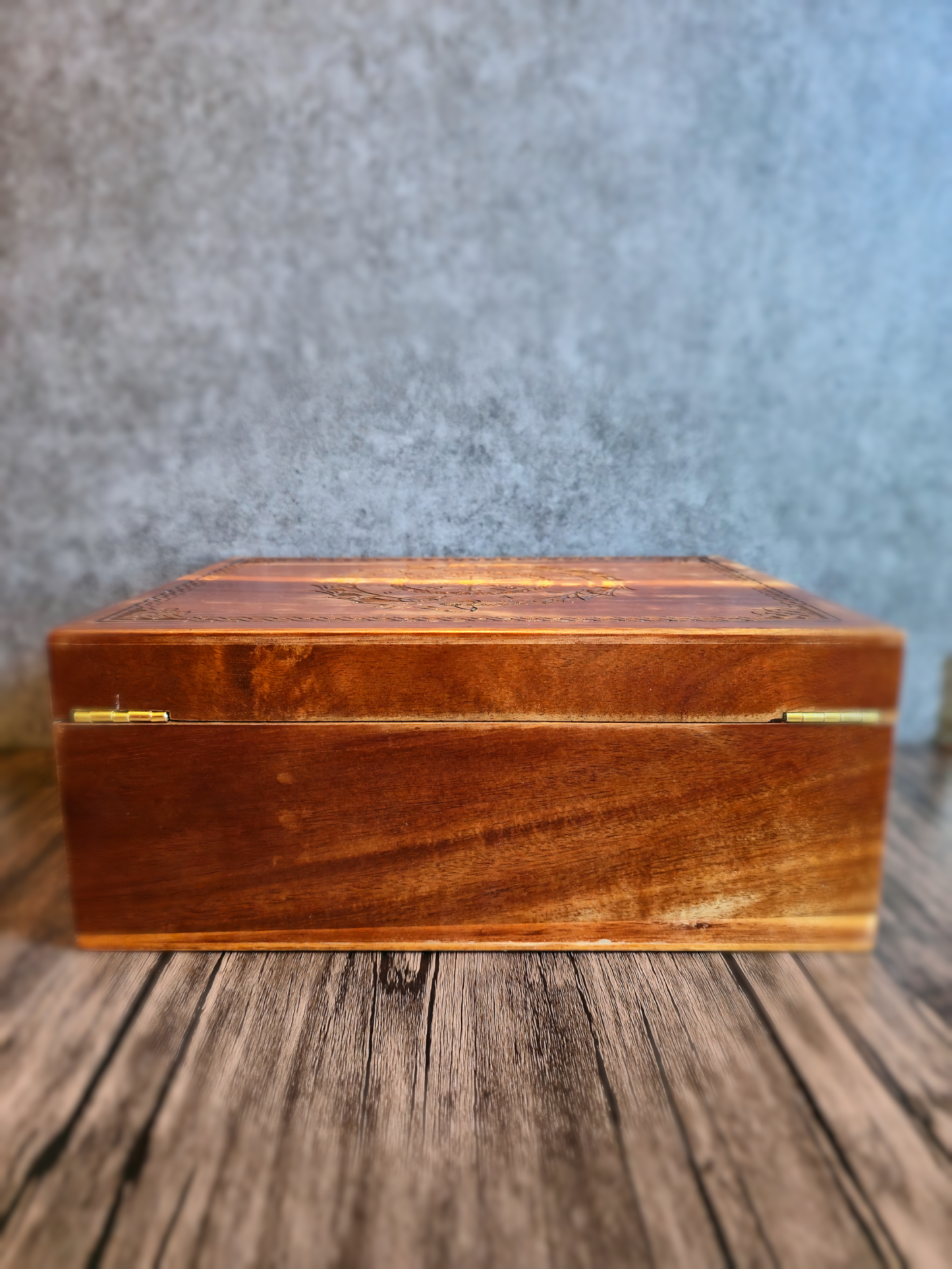 A low-angle side-on view of the handcrafted wooden box with its lid closed, revealing smooth brown and amber wood grain on the front panel and the subtle edge of the intricately carved Norse dragon design on the top, all set against a soft gray background.
