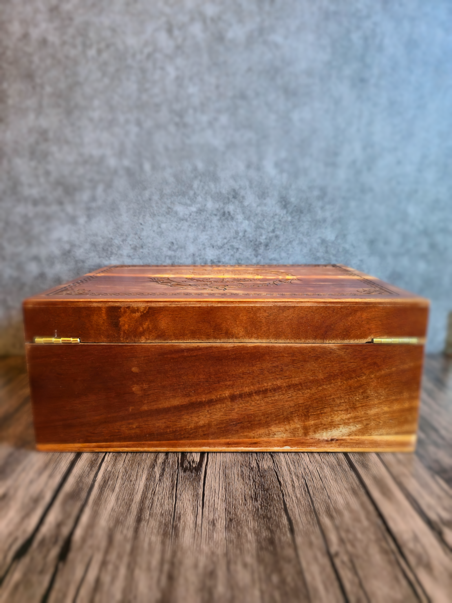 A low-angle side-on view of the handcrafted wooden box with its lid closed, revealing smooth brown and amber wood grain on the front panel and the subtle edge of the intricately carved Norse dragon design on the top, all set against a soft gray background.
