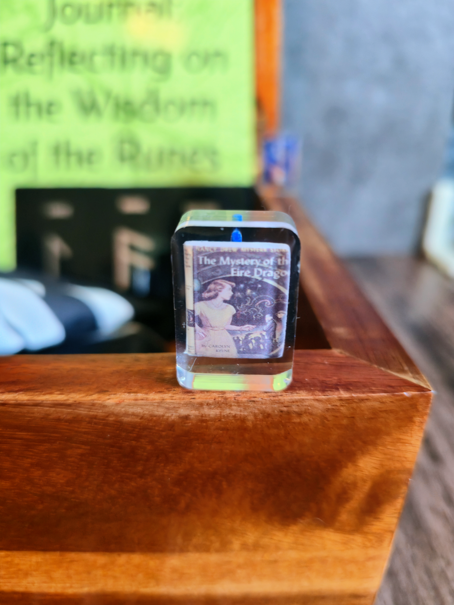 Small book or magazine inside a transparent box on a wooden surface with a blurred background.