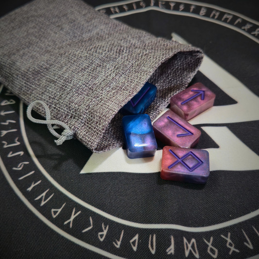 A gray linen drawstring pouch spilling hand-poured resin runestones in swirling blue, purple, and red hues, each etched with an Elder Futhark symbol, set atop a black cloth printed with a white circular runic border and triangular Norse symbol.