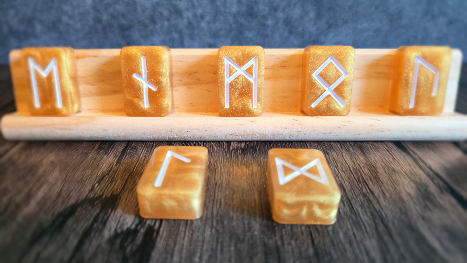 Gold runestones with white symbols standing upon a birchwood stand with a grey background and wooden table