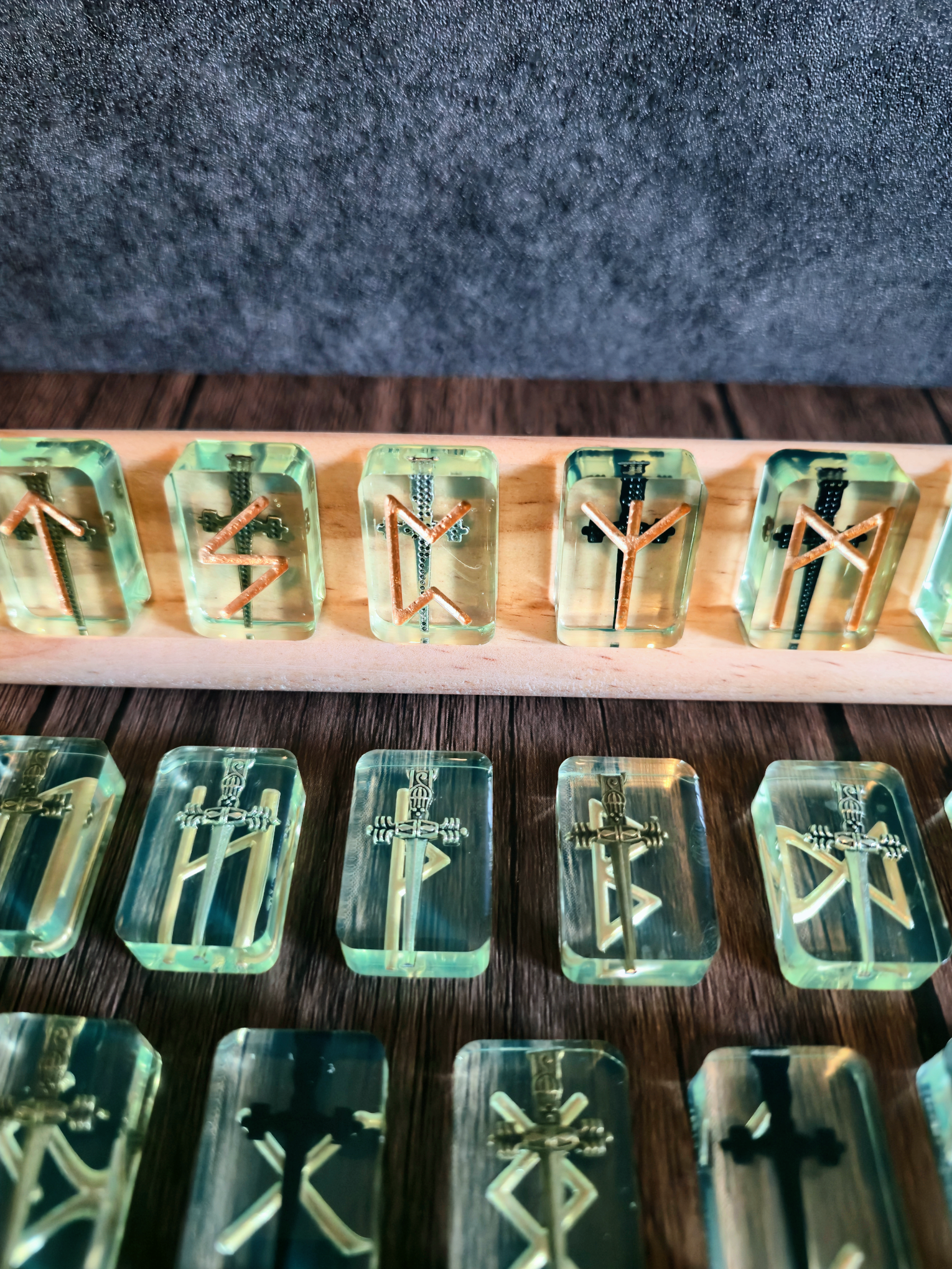 Greenish clear runestones with swords embedded with gold runic symbols on birchwood stand with grey background on wooden table.