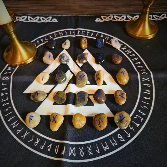 Twenty-four smooth tumbled stones in shades of beige, green, brown, and black, each hand-painted with a bright blue Elder Futhark rune, are arranged in a stacked triangle pattern atop a black cloth printed with a large white Valknut symbol encircled by runic text. Two brass candle holders with ivory tapers flank the display on a wooden surface.