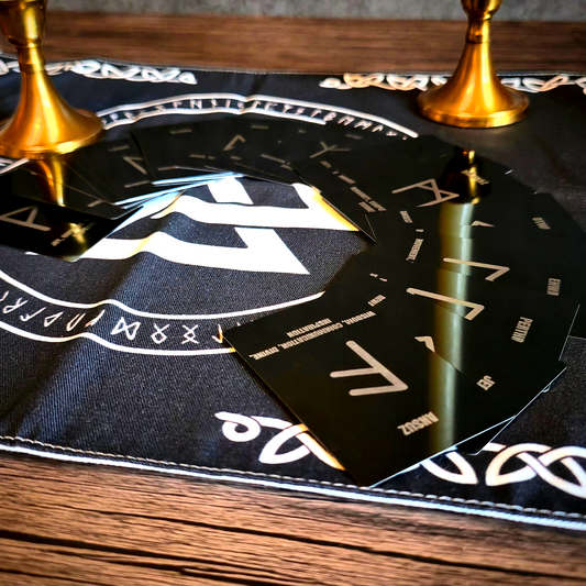 A semi-circle of matte black rune cards, each emblazoned with a white Norse rune glyph and its name (e.g., ‘Perthro,’ ‘Ansuz’) in small text, fanned across a black woven cloth bearing a central white Valknut and ring of runic script. Two brass candle holders with ivory tapers flank the display on a wooden table.