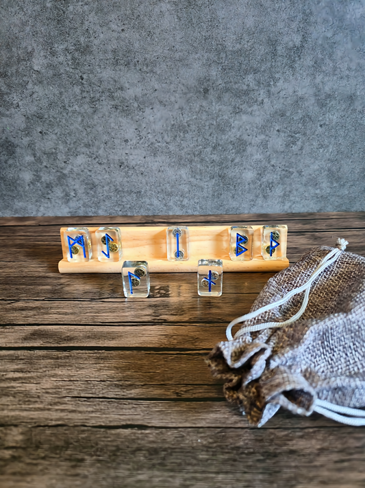 Clear resin runestones etched with blue Norse runes displayed on a light wood stand, next to a textured burlap pouch on a weathered wooden table against a soft gray backdrop.