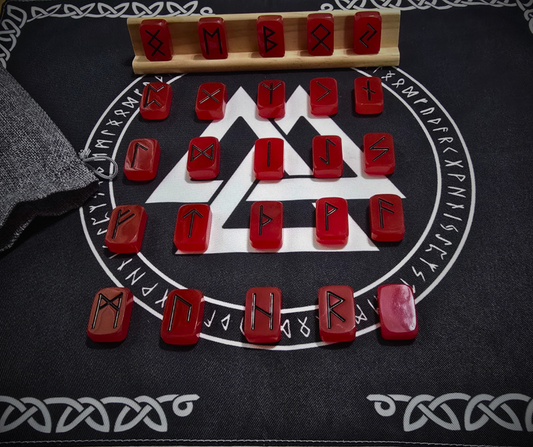 Viking Fire Runestone Set: 24 glossy red resin Elder Futhark runes with bold black symbols, arranged in a circular spread on a black rune cloth printed with a white Valknut and runic border, complete with a gray linen pouch and birch wood display stand.