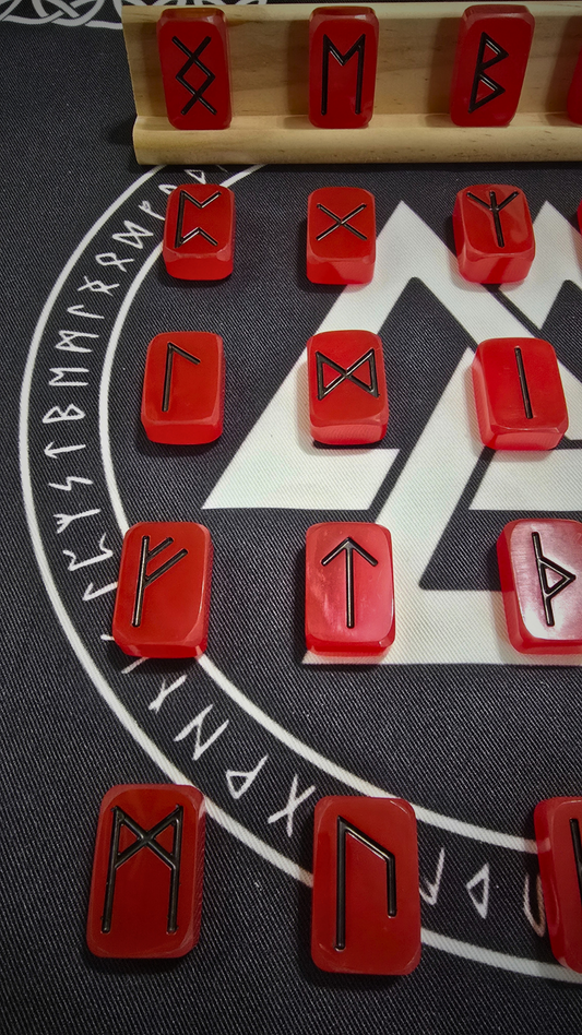 Viking Fire Runestone Set: glossy red resin runes engraved with bold black Elder Futhark symbols, arranged in neat rows on a black cloth printed with a white Valknut and circular runic border, with five runes showcased on a birch wood display rack above.