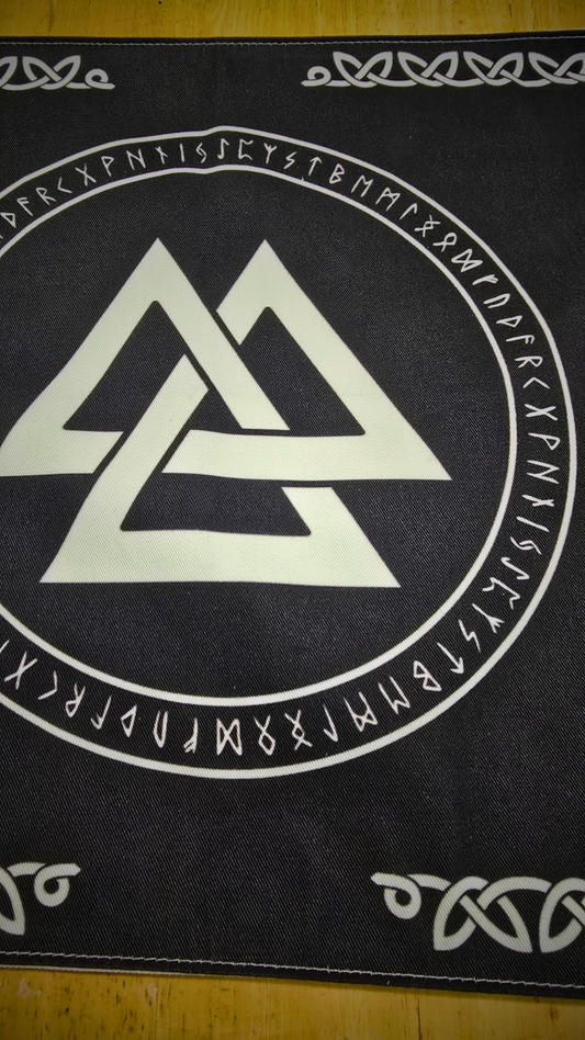 Black divination mat printed with a bold white Valknut interlocking-triangles symbol at center, surrounded by a circular Elder Futhark rune inscription and framed with Celtic knotwork trim on a wooden tabletop.